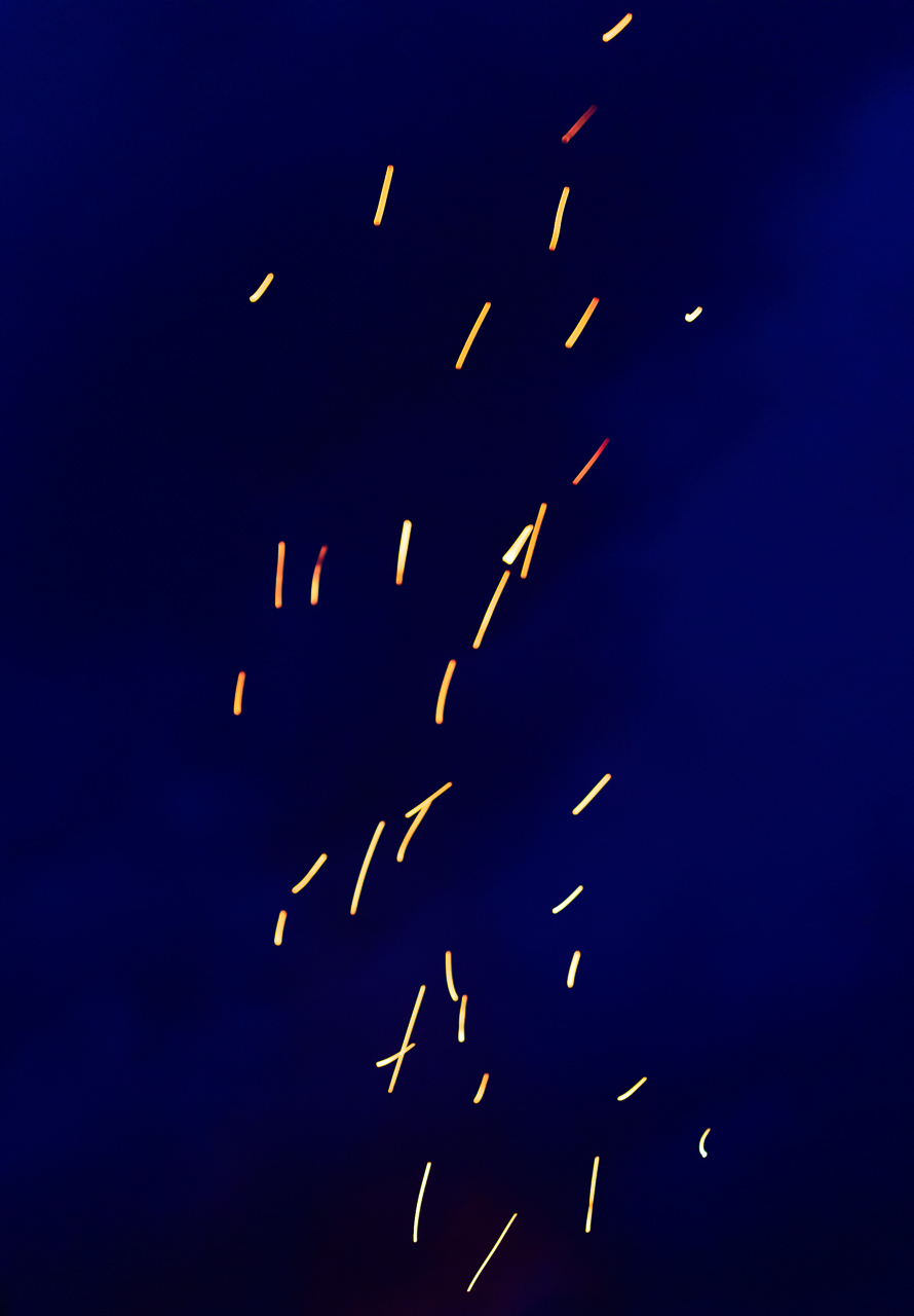 Fliegende Funken eines Lagerfeuers wirken vor einem schwarzblauen Nachthimmel durch eine längere Belichtungszeit wie Pinselstriche in einem abstrakten Gemälde.