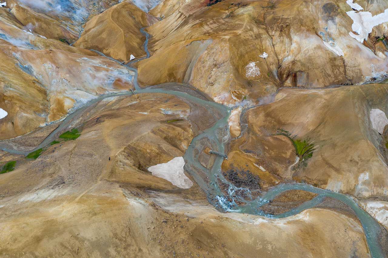 Aus der Vogelperspektive gesehen: das Geothermalgebiet Kerlingarfjöll in Island. Pastellfarbene bunte Berge, dampfende heiße Quellen und Schneefelder kennzeichnen die Landschaft. Zwei Wanderer im Bild links markieren die Größenverhältnisse.