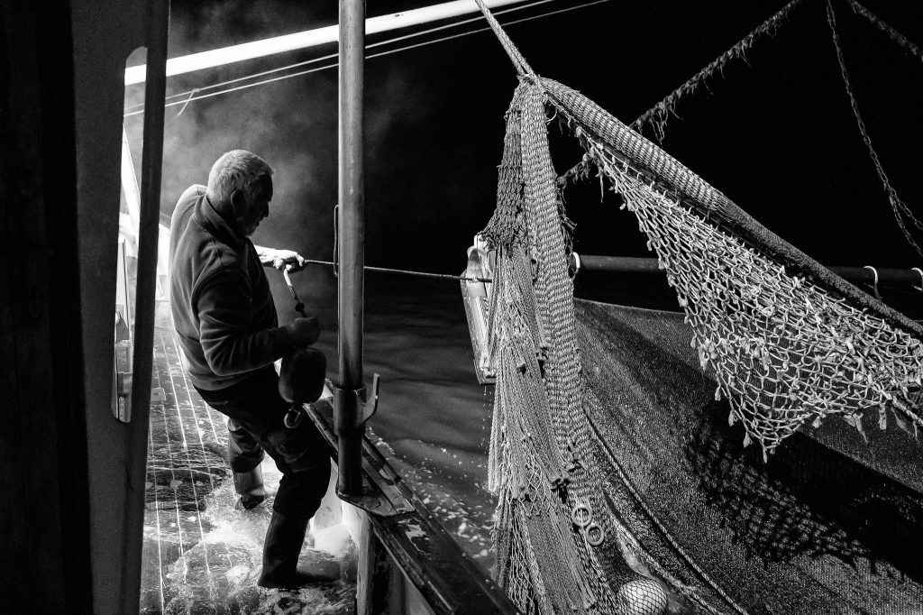 Beim Einholen der Netze – hier durch den Krabbenkutter-Kapitän Fritz Ronnebeck an Bord der Annemarie – treffen Licht, Wasser und Dampf aufeinander. Die nächtliche Arbeit an Deck ist körperlich anspruchsvoll und folgt einem präzisen Ablauf, der sich über Jahrzehnte kaum verändert hat.